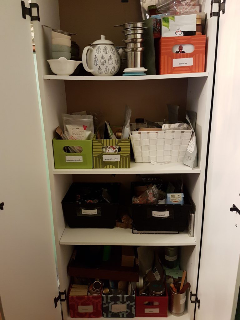 A white plywood cabinet with four shelves inside. Each shelf contains several boxes of loose-leaf tea, and each box belongs to its own category.
