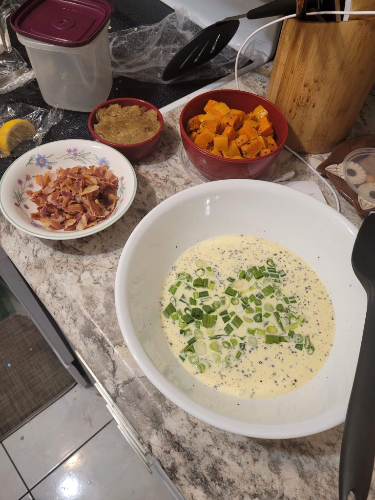 A variety of pie filling ingredients before mixing: a white bowl filled with cream, eggs and green onions; a small bowl of crumbled bacon, and small containers of caramelized onions and roasted squash.