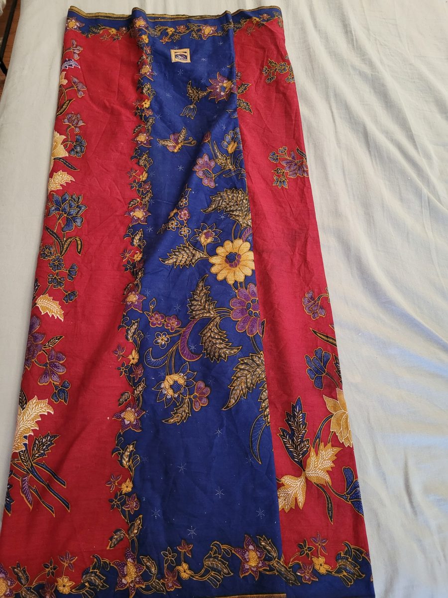 A bolt of cloth sitting on a bedspread. The cloth is crimson and navy blue, and covered in multicoloured flowers.